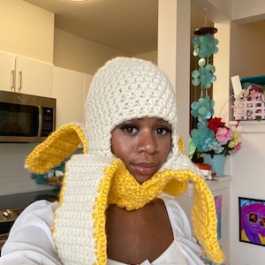 Peut inclure: Ensemble fait main composé d'un bonnet et d'une écharpe au crochet. Le bonnet est de couleur crème, l'écharpe a des bords jaunes et est conçue pour ressembler à une peau de banane. L'ensemble est porté sur un chemisier blanc.