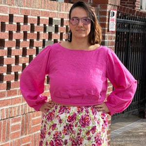 May include: A bright pink long-sleeved cropped top with a wide neckline and gathered sleeves paired with white trousers featuring a vibrant floral pattern of pink and purple roses with green leaves. The outfit is worn with tinted aviator sunglasses.
