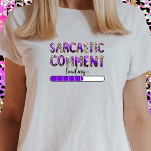 May include: White t-shirt featuring the text "SARCASTIC COMMENT loading..." in purple and gold lettering. A purple loading bar graphic is below the text. The shirt is a casual, everyday wear item.