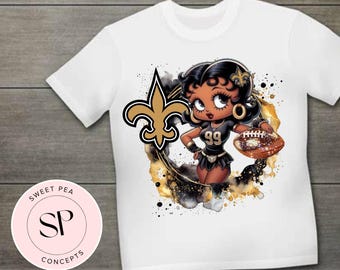 May include: White t-shirt with a cartoon image of a woman wearing a football uniform with the number 99. The woman is holding a football and is surrounded by gold and black paint splatters. The image also includes a gold fleur-de-lis.