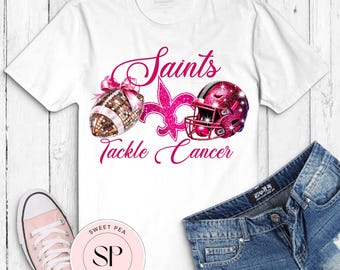 May include: White t-shirt featuring a pink design with a football, helmet, and fleur-de-lis. The text "Saints Tackle Cancer" is written in pink script. A pair of denim shorts and pink sneakers are also visible.