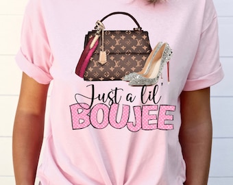 May include: Pink t-shirt with a graphic of a brown and gold handbag and a silver sparkly high heel shoe. The text "Just a lil' BOUJEE" is printed in pink with white polka dots.