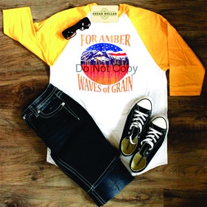 May include: A white and yellow baseball tee shirt with a graphic of a mountain range and an American flag with the text "For Amber Waves of Grain". The shirt is paired with blue jeans and black sneakers.