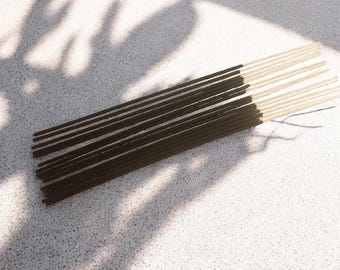May include: A collection of thin incense sticks featuring dark brown, textured coating on the upper portions and smooth, light beige wooden bases. The sticks are arranged in a parallel cluster, showcasing the contrast between the dark aromatic material and the natural wood ends, highlighting the simple, slender cylindrical form of each individual incense stick.