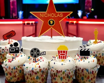 May include: A white cake with a red star on top that says "NICHOLAS". The cake is surrounded by cupcakes with various movie-themed decorations, including film reels, clapperboards, and popcorn.