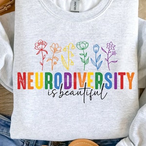 Peut inclure: Un sweat-shirt gris avec un motif arc-en-ciel qui dit "Neurodiversity is beautiful". Le motif représente une rangée de fleurs stylisées.
