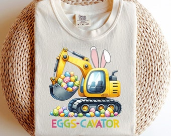 May include: A white t-shirt with a yellow cartoon excavator with bunny ears. The excavator is holding a scoop full of colorful Easter eggs. The text "EGGS-CAVATOR" is printed below the excavator.