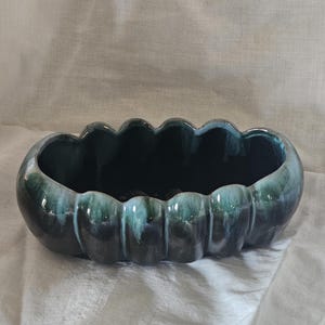 May include: A ceramic planter with a scalloped rim and a glossy, dark teal glaze. The oval-shaped container has a textured surface and a deep interior, suitable for holding plants or other decorative items.