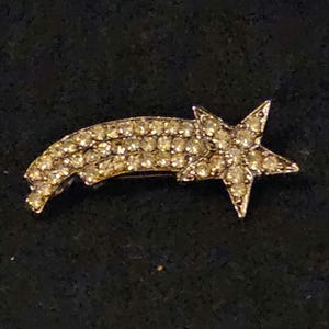 May include: A vintage brooch in the shape of a shooting star. The brooch is covered in small, sparkling rhinestones, with a star at the end of the tail. The brooch is set against a dark background.
