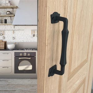 May include: Black metal door handle with a decorative design. The handle is mounted on a light wood door.