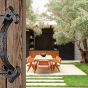 May include: Black cast iron door handle with a curved design. The handle is mounted on a wooden door with screws. The handle is in focus, while the background is blurred and shows a patio with a table and chairs.