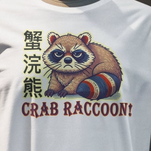 May include: White t-shirt with a cartoon illustration of a raccoon with a crab leg for a tail. The text "蟹 浣 熊" is written in Japanese above the raccoon and "CRAB RACCOON!" is written below the raccoon.