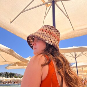 May include: A crocheted sun hat in brown, orange, and beige. The hat has a wide brim and a tag that reads "baggies". The person is wearing an orange swimsuit. The background includes a blue sky and beach umbrellas.