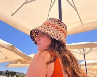May include: A crocheted sun hat in shades of brown, orange, and beige. The hat has a wide brim and a tag that reads "baggies". The person is wearing an orange swimsuit. The background includes a blue sky and beach umbrellas.