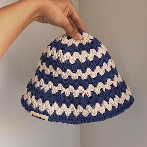 Puede incluir: Un sombrero de cubo de ganchillo azul y crema. El sombrero presenta un patr&oacute;n repetitivo de cuadrados azules y crema. El ala es ligeramente acampanada. La etiqueta del sombrero dice "baddles".
