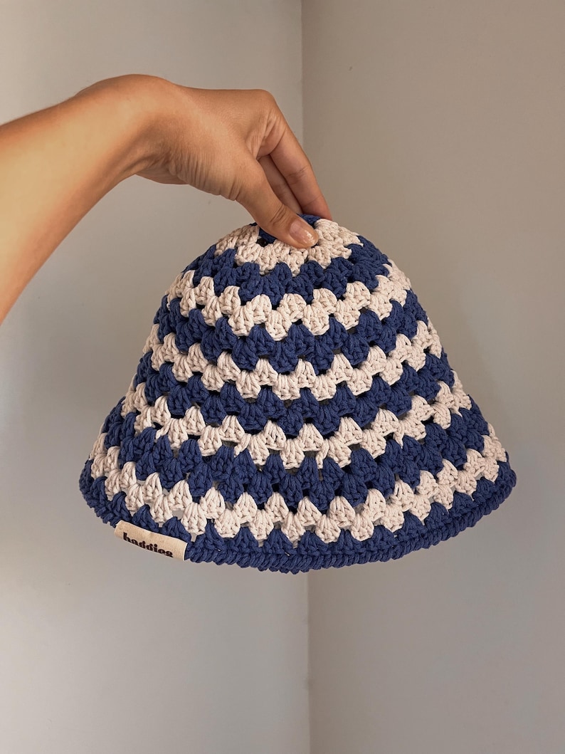 Puede incluir: Un sombrero de cubo de ganchillo azul y crema. El sombrero presenta un patr&oacute;n repetitivo de cuadrados azules y crema. El ala es ligeramente acampanada. La etiqueta del sombrero dice "baddles".