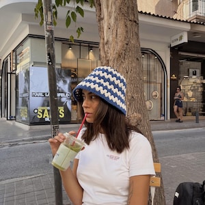 Könnte beinhalten: Ein weißes T-Shirt mit rotem "GUESS JEANS"-Aufdruck, getragen mit einem blau-weiß gehäkelten Fischerhut. Die Person trinkt ein grünes Getränk mit einem roten Strohhalm. Im Hintergrund sind ein Baum, ein Schaufenster und der Text "SUMMER SALES" zu sehen.