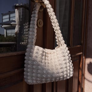 May include: Cream-colored, hand-crocheted tote bag with a textured, bobble stitch design. The bag features a thick, sturdy strap and is hanging on a wooden door. The bag's neutral color and unique texture make it a stylish accessory.