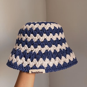 Puede incluir: Un sombrero de pescador de ganchillo azul, blanco y beige. El sombrero presenta un patr&oacute;n geom&eacute;trico y una peque&ntilde;a etiqueta que dice "baddies". El sombrero est&aacute; hecho a mano y tiene un aspecto suave y texturizado.
