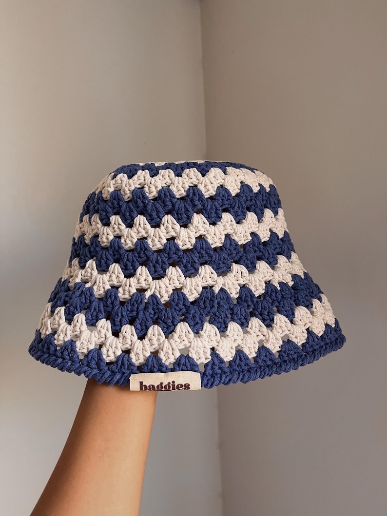 Puede incluir: Un sombrero de pescador de ganchillo azul, blanco y beige. El sombrero presenta un patr&oacute;n geom&eacute;trico y una peque&ntilde;a etiqueta que dice "baddies". El sombrero est&aacute; hecho a mano y tiene un aspecto suave y texturizado.