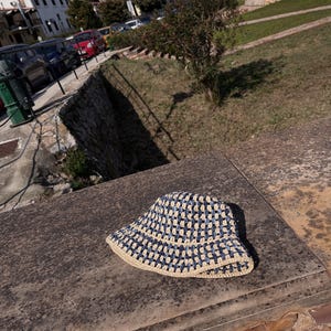May include: A blue and beige crochet bucket hat. The hat features a square pattern in navy blue and beige. The brim is beige. The hat is sitting on a weathered stone surface.