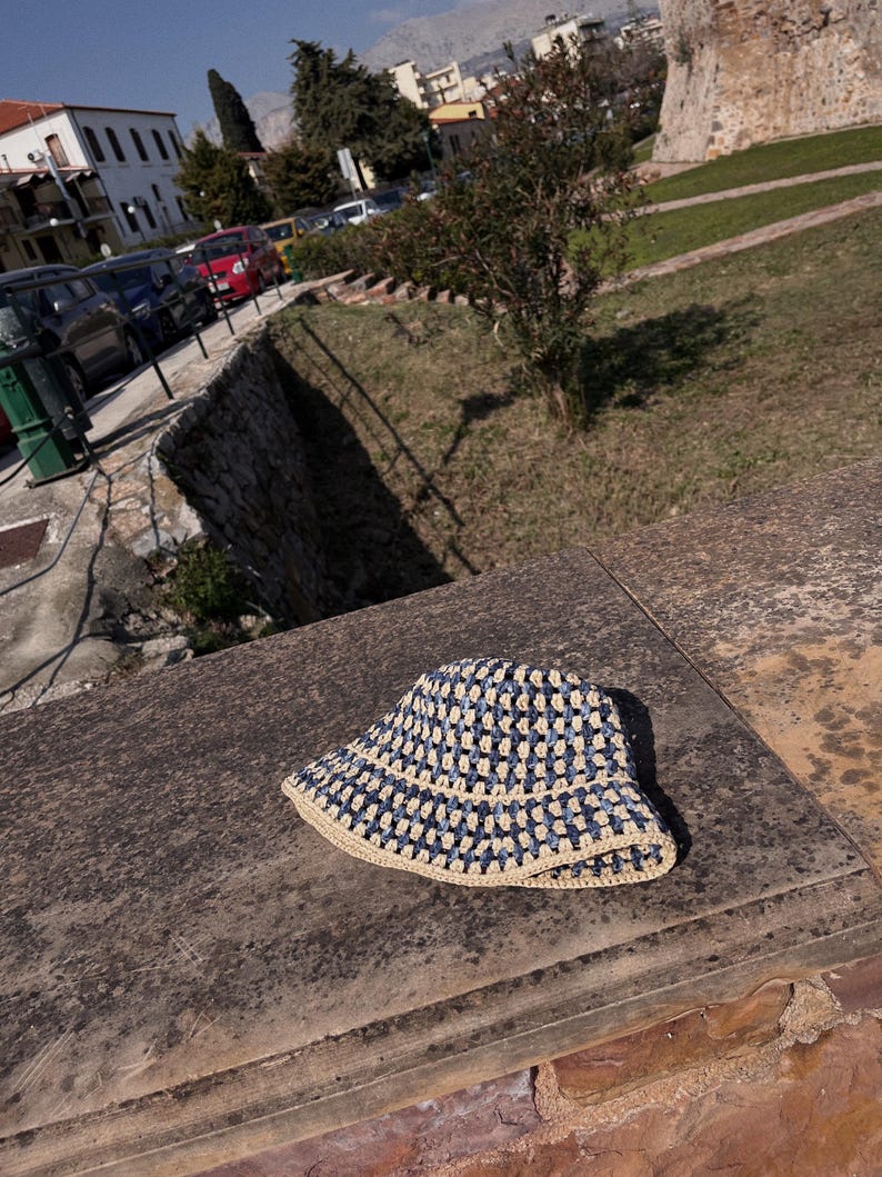 May include: A blue and beige crochet bucket hat. The hat features a square pattern in navy blue and beige. The brim is beige. The hat is sitting on a weathered stone surface.