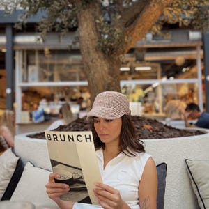 Puede incluir: Una mujer con un sombrero de ganchillo beige y una camisa sin mangas blanca lee un menú que dice "BRUNCH". La escena está ambientada al aire libre, con un gran árbol al fondo y otras personas visibles. El menú es de color crema con texto negro.