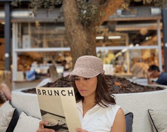 Może przedstawiać: Kobieta w beżowym kapeluszu z szydełka i białej koszulce bez rękawów czyta menu z napisem "BRUNCH". Scena rozgrywa się na zewnątrz, z dużym drzewem w tle i innymi widocznymi osobami. Menu jest kremowe z czarnym tekstem.