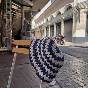 Puede incluir: Un gorro de ganchillo azul y crema con un patr&oacute;n geom&eacute;trico descansa sobre una silla blanca. El gorro hecho a mano presenta un dise&ntilde;o repetitivo de cuadrados azules y crema. El gorro tiene la palabra "baddies".