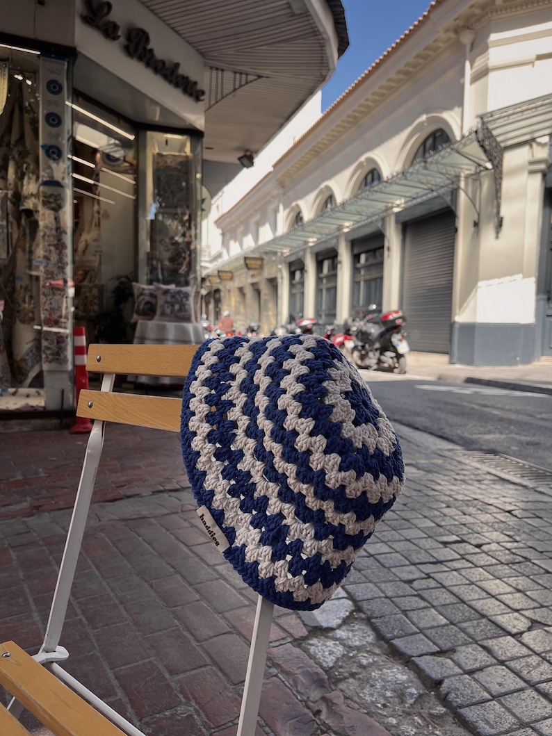 Puede incluir: Un gorro de ganchillo azul y crema con un patr&oacute;n geom&eacute;trico descansa sobre una silla blanca. El gorro hecho a mano presenta un dise&ntilde;o repetitivo de cuadrados azules y crema. El gorro tiene la palabra "baddies".