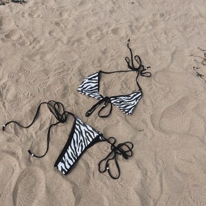 Puede incluir: Un bikini de estampado de cebra en blanco y negro con lazos negros. La parte superior del bikini es de estilo halter y la parte inferior es de estilo atrevido.