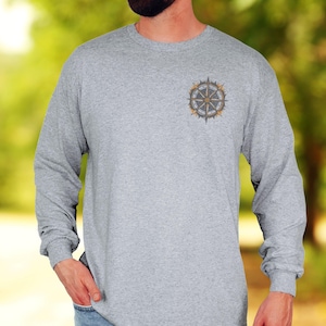 May include: A heather gray long-sleeve shirt featuring a small, circular compass graphic printed on the upper left chest. The shirt has a crew neckline and long sleeves, paired with light blue denim jeans, creating a casual look.