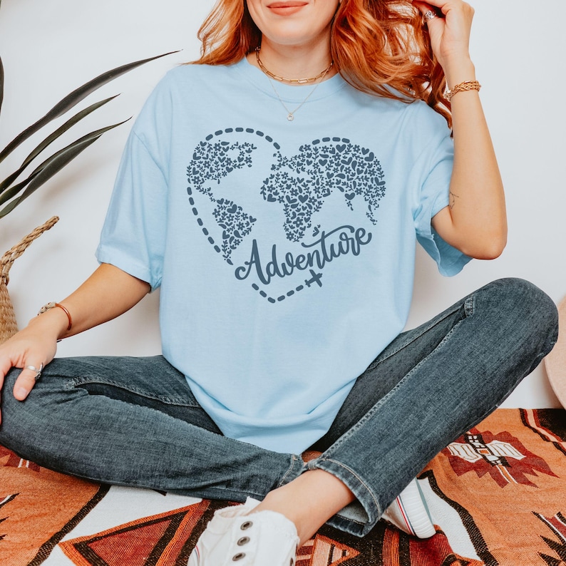 May include: Light blue t-shirt featuring a heart-shaped world map design filled with floral and leaf patterns. The word "Adventure" is written in cursive below the map, with a dotted line and airplane graphic. The shirt is made of soft material.