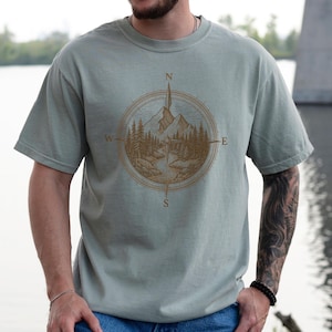 Peut inclure: T-shirt vert sauge avec un motif de boussole détaillé représentant une scène de montagne et de forêt. La rose des vents indique les points cardinaux. Le t-shirt est de style décontracté et confortable, pour les amateurs de plein air et de nature.