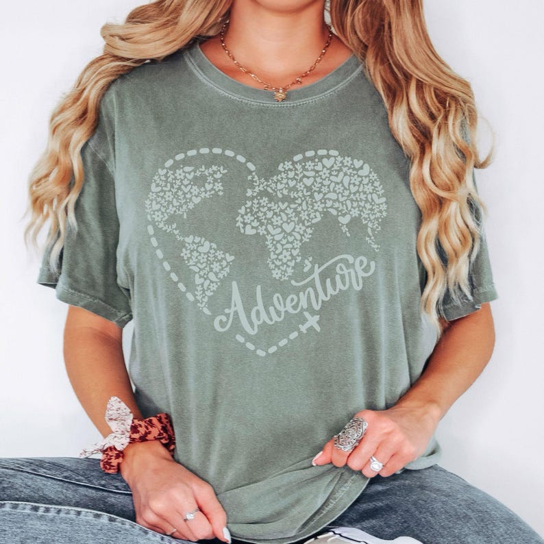 May include: Sage green t-shirt featuring a heart-shaped world map design filled with small hearts. The word "Adventure" is written in cursive below the map. A dotted line with a small airplane graphic surrounds the map.
