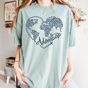 May include: A light teal t-shirt featuring a heart-shaped world map design filled with floral patterns. The word "Adventure" is written in cursive below the map, with a dotted line and airplane graphic. The shirt is a soft, comfortable material.
