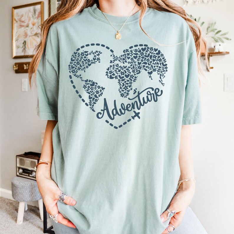 May include: A light teal t-shirt featuring a heart-shaped world map design filled with floral patterns. The word "Adventure" is written in cursive below the map, with a dotted line and airplane graphic. The shirt is a soft, comfortable material.