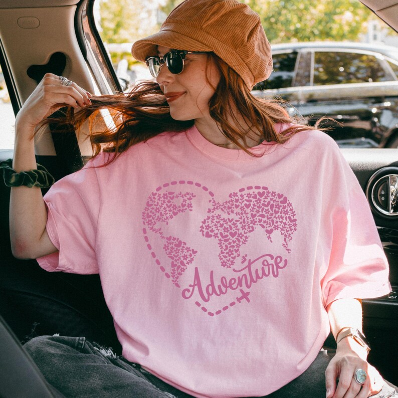 May include: A light pink t-shirt featuring a heart-shaped world map design filled with floral patterns. The word "Adventure" is written in a cursive font within the heart. The shirt is a soft, casual style.