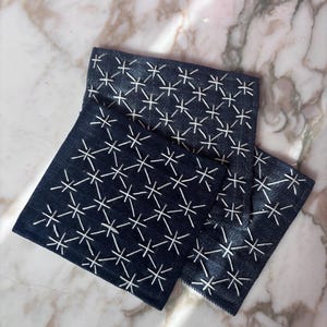 May include: Three navy blue square coasters with a white embroidered starburst pattern. The coasters are made of a textured fabric and are arranged on a marble surface. The coasters are a decorative home accessory.