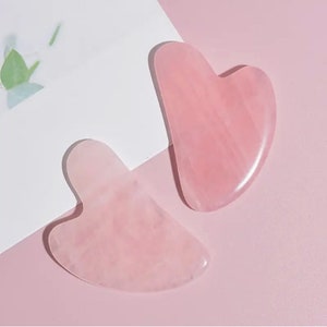 May include: Two pink heart-shaped rose quartz gua sha tools. The tools are made of smooth, polished rose quartz and have a slightly curved shape.