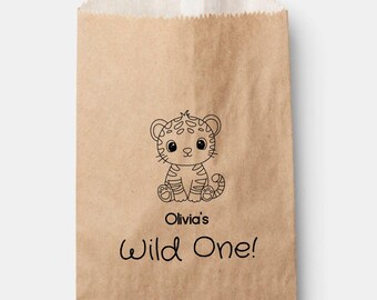 May include: A brown paper gift bag featuring a black and white illustration of a cartoon tiger cub. The text "Olivia's Wild One!" is printed below the tiger. The bag has a folded top edge.