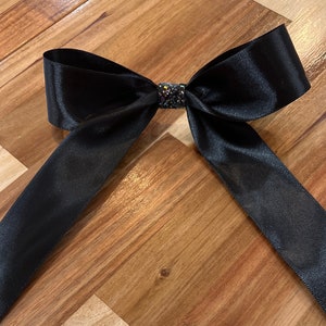 May include: A black satin bow with a sparkly center. The bow is tied with long tails and is lying on a wooden surface.