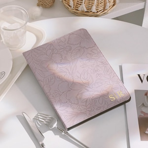 May include: A light purple journal with a floral embossed design and gold initials "S R". The journal is on a white table with silverware, a glass, and a decorative basket. A magazine is visible in the background.