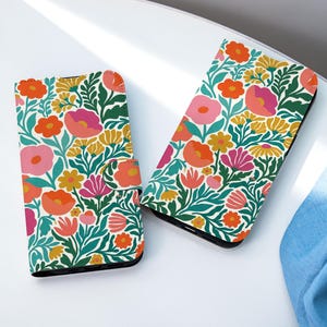 Könnte beinhalten: Zwei Handyhüllen mit floralem Muster mit einem lebendigen Muster aus orangefarbenen, rosa und gelben Blumen und grünen Blättern auf weißem Hintergrund. Eine Hülle ist offen, die andere geschlossen. Die Hüllen liegen auf einer weißen Oberfläche.