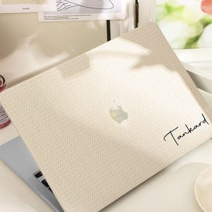 Könnte beinhalten: Eine beige Laptop-Hülle mit einem strukturierten Stoffdesign. Auf der Hülle steht das Wort "Tankard" in Handschrift. Das Apple-Logo ist sichtbar. Der Laptop steht auf einem weißen Schreibtisch mit einer rosa Parfümflasche und einer Teetasse.