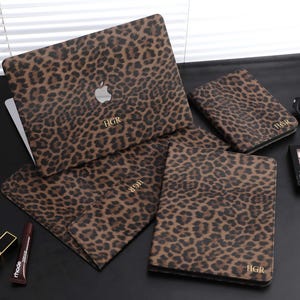 Puede incluir: Un conjunto de accesorios con estampado de leopardo, que incluye una funda para portátil, una funda para tableta y varias otras fundas. Cada artículo presenta un patrón de manchas marrones y negras y las iniciales "HGR" en dorado. Sobre una superficie negra.