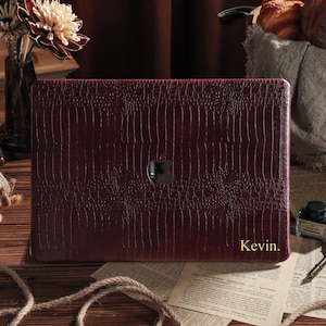 Könnte beinhalten: Eine weinrote Laptop-Hülle mit einem strukturierten, krokodilartigen Muster. Die Hülle hat ein schwarzes Apple-Logo und den Namen "Kevin" in goldfarbenen Buchstaben. Für einen Laptop.