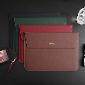 Może przedstawiać: Skórzane etui na laptopa w kolorze czarnym, zielonym, czerwonym i brązowym, z napisem "Robin" w kolorze złotym na brązowym etui. Na zdjęciu znajduje się również paleta cieni do powiek, błyszczyk do ust, szklanka wody i okulary przeciwsłoneczne.