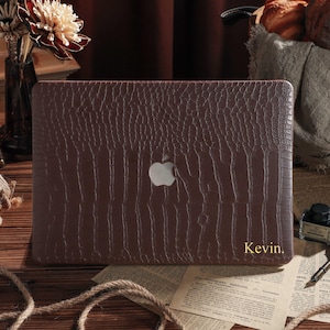 Könnte beinhalten: Eine braune Laptop-Hülle mit strukturiertem Muster und dem Apple-Logo. Der Name "Kevin" ist goldfarben unten rechts aufgedruckt. Die Hülle liegt auf einer Holzoberfläche mit Accessoires im Vintage-Stil.