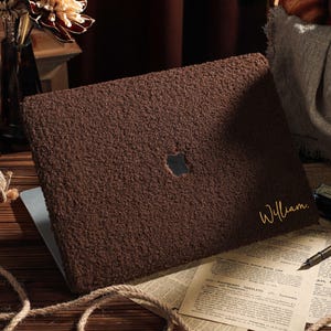 Può includere: Una custodia per laptop marrone con una superficie strutturata. La custodia ha un ritaglio per il logo Apple e il nome "William" in corsivo dorato. Il laptop è aperto, rivelando uno schermo argentato.